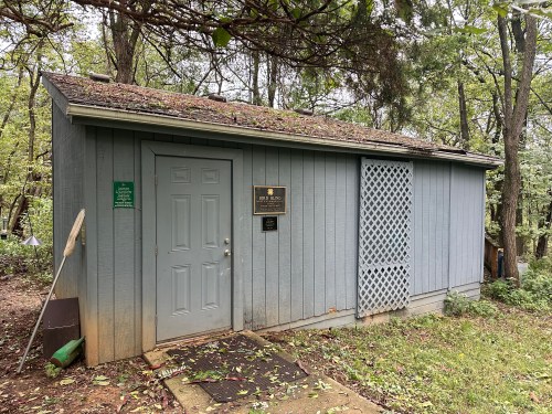 A small, gray wooden building with a door, plaque, and lattice window, surrounded by trees and greenery.