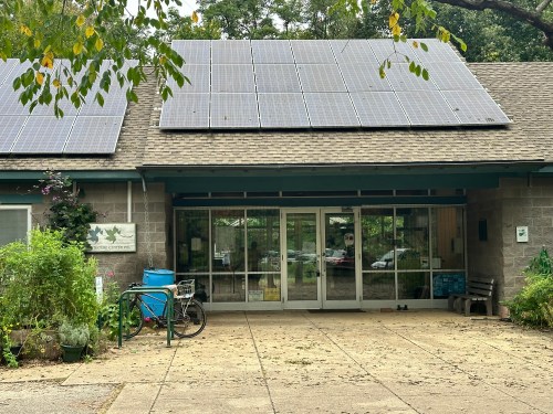 A building with solar panels on the roof, featuring a glass entrance and surrounded by greenery and plants.