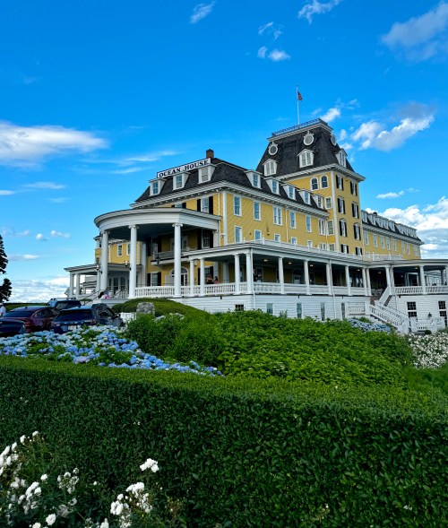 A large yellow hotel with a grand porch, surrounded by green bushes and flowers, under a blue sky with clouds.
