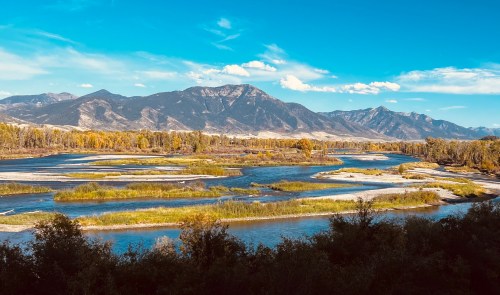 A scenic view of a river winding through green and golden landscapes, framed by mountains under a clear blue sky.