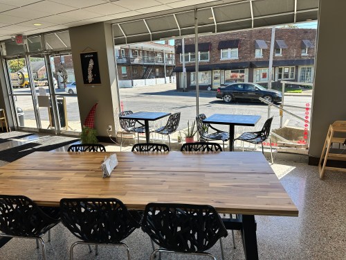 A spacious café interior with a large wooden table, black chairs, and a view of the street through large windows.