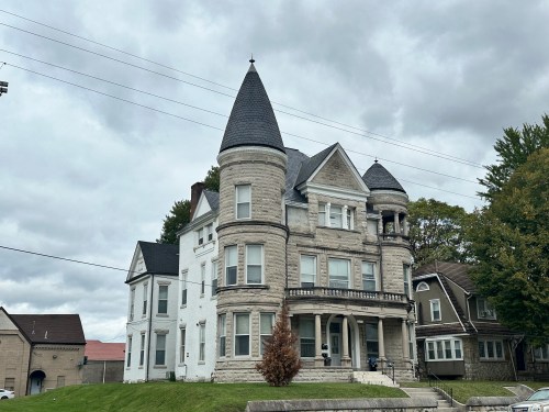 A large, historic house with a turret, gray stone exterior, and multiple gables, set against a cloudy sky.