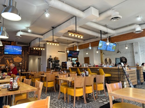 A cozy restaurant interior with wooden tables, yellow chairs, and a coffee station, featuring TVs and decorative lighting.