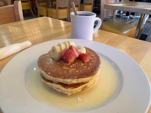 A stack of pancakes topped with banana slices and strawberries, served with syrup and a cup of coffee in the background.