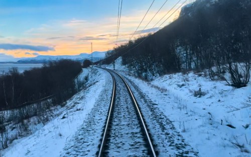 A winding train track through a snowy landscape at sunset, with mountains in the background and a colorful sky.