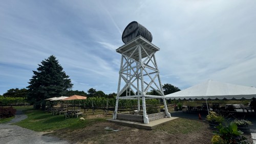 A wooden water tower stands beside a vineyard, with picnic tables and a large tent in the background under a blue sky.