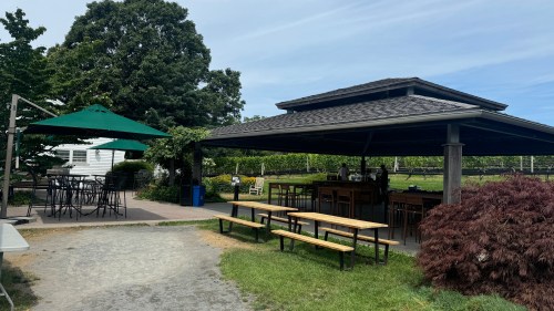 Outdoor seating area with picnic tables and a covered bar, surrounded by greenery and a clear blue sky.