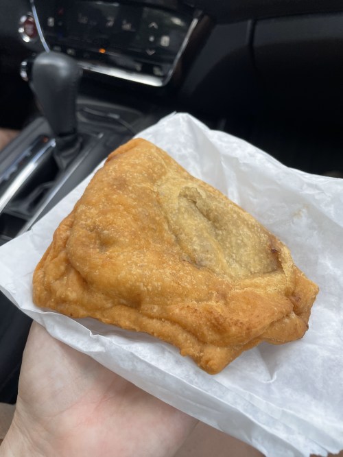 A hand holding a golden-brown fried pastry wrapped in white paper, with a car interior in the background.