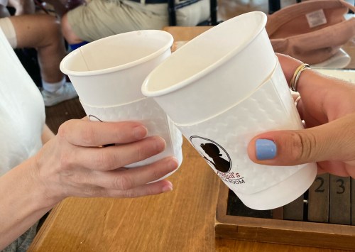 Two hands clink white cups together, celebrating a moment, with a wooden table in the background.