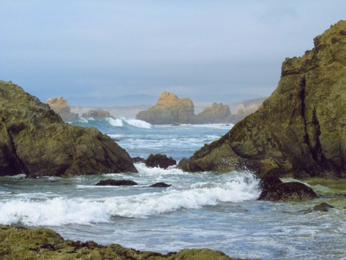 A serene coastal scene with rocky cliffs, gentle waves, and misty mountains in the background under a cloudy sky.