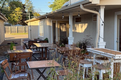 Outdoor seating area with wooden tables and chairs, surrounded by plants and string lights, next to a building.