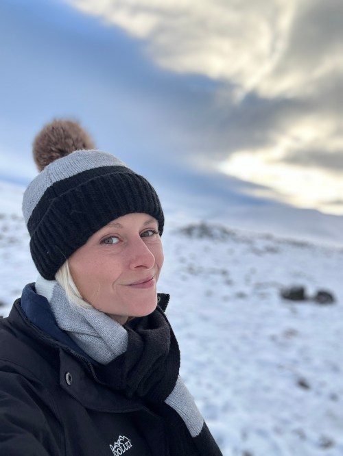 A woman in a winter hat and scarf smiles in a snowy landscape under a cloudy sky.