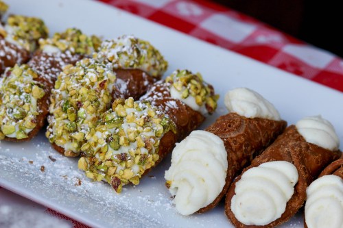 A plate of cannoli filled with cream, one topped with crushed pistachios and the other with a smooth white frosting.