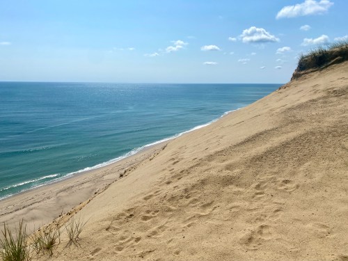 A sandy dune slopes down to a calm ocean under a clear blue sky with a few clouds.