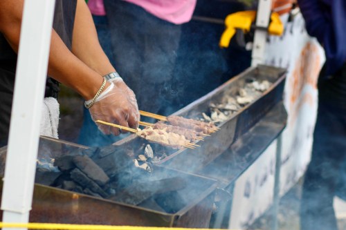 A person wearing gloves grills skewers of meat over a smoky barbecue, with charcoal visible in the background.