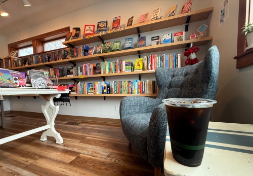 A cozy reading nook with a gray chair, a table with a drink, and a bookshelf filled with colorful books.