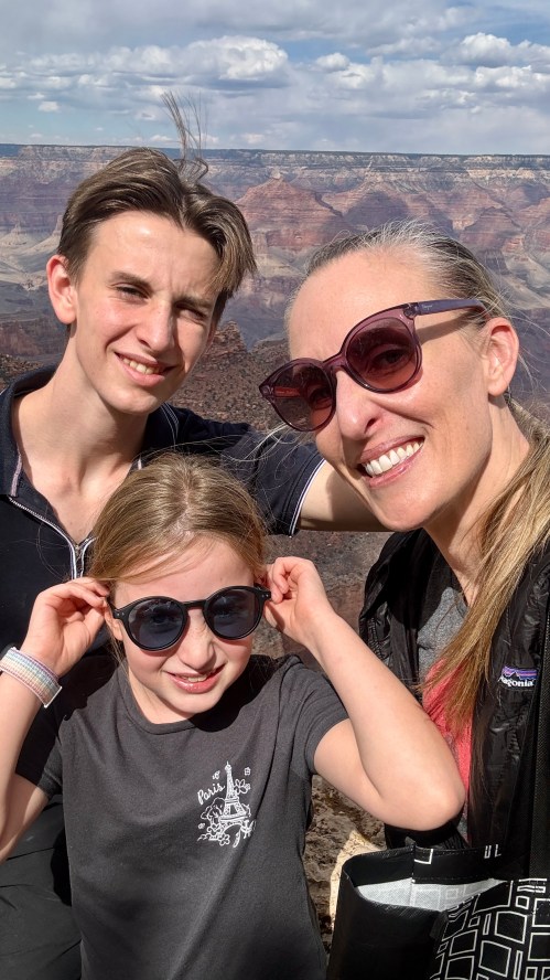 A woman and two children wearing sunglasses smile for a selfie at the Grand Canyon, with a scenic view in the background.