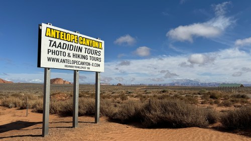 A sign for Antelope Canyon tours stands in a desert landscape under a blue sky with scattered clouds.