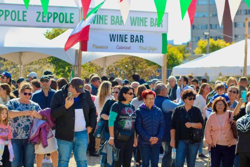 A crowd of people at an outdoor festival with tents for wine and zeppole, decorated with colorful banners.