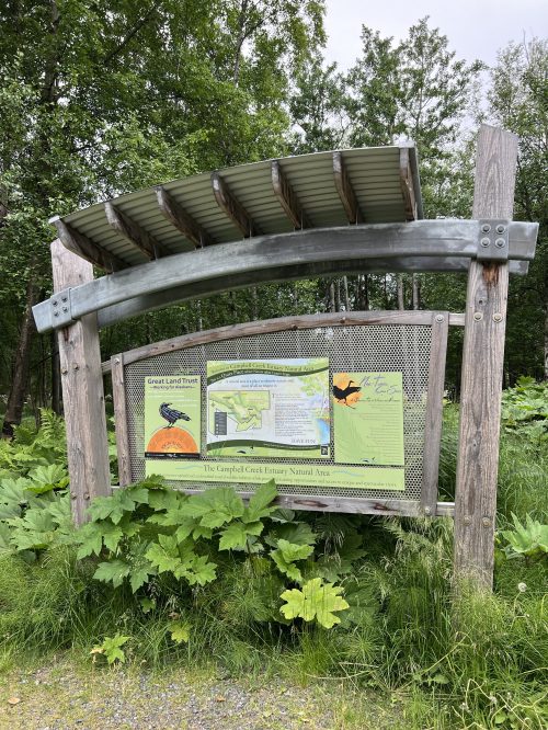 Sign at Campbell Creek Estuary Natural Area, featuring a map and information about the local environment, surrounded by greenery.