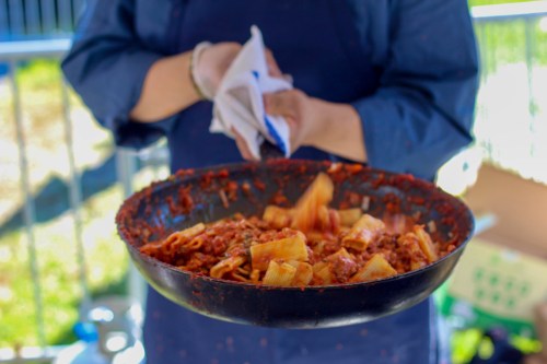 A person holds a large pan of pasta in a rich red sauce, with a towel in their other hand, ready to serve.