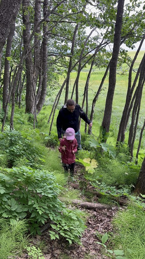 A woman and a child walk down a wooded path surrounded by lush greenery and trees.