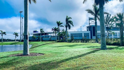 A modern sports facility surrounded by palm trees and a pond, under a partly cloudy sky.