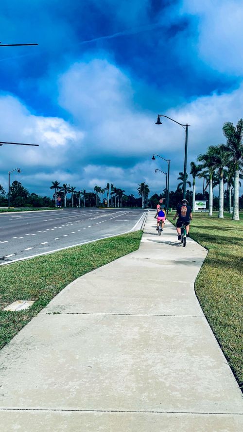 Two children ride bicycles along a wide sidewalk beside a road lined with palm trees under a cloudy sky.