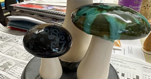 Three decorative mushroom sculptures in varying sizes and colors, displayed on a table with papers and objects in the background.