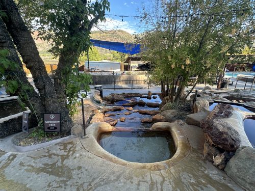 A serene outdoor spa area featuring a natural stone pathway, water features, and lush greenery under a clear blue sky.