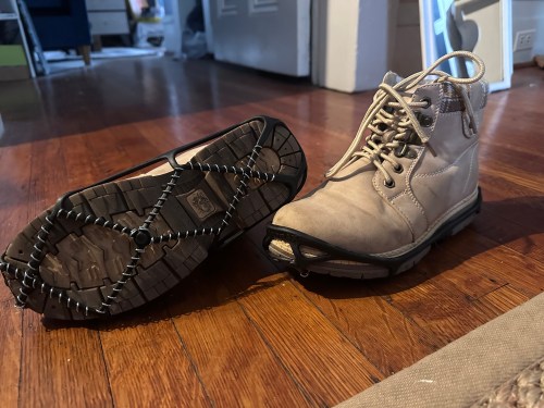 A pair of light-colored boots, one with a traction device attached, on a wooden floor.