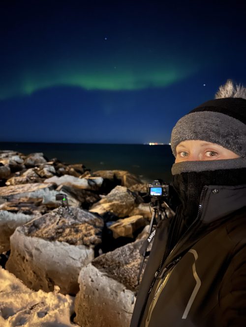 A person in winter clothing stands by rocky shore at night, with the northern lights glowing in the sky above.