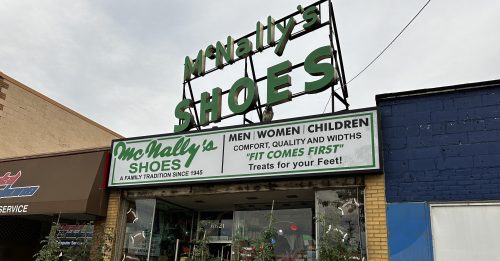 A storefront for McNally's Shoes, featuring a sign and window displays with plants and shoes for men, women, and children.