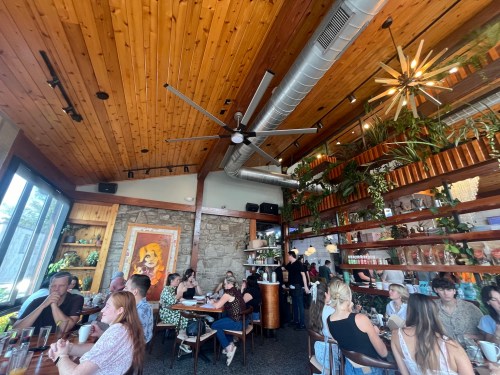 A lively restaurant interior with wooden beams, plants, and patrons enjoying their meals at various tables.
