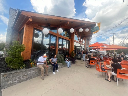 A modern restaurant with a wooden exterior, outdoor seating, and people enjoying their meals on a sunny day.