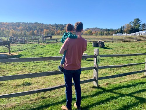 A person holds a child while looking at a scenic farm with green fields and trees in the background.