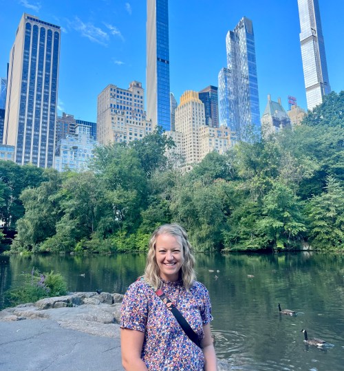A smiling woman stands by a serene pond, surrounded by lush greenery and tall city skyscrapers in the background.