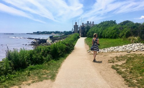 A woman walks along a coastal path with lush greenery and a view of the ocean under a blue sky.