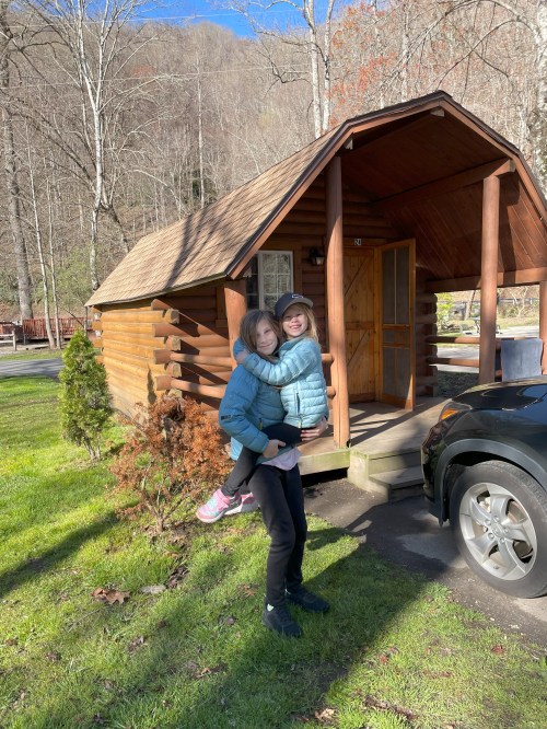 Two girls stand outside a wooden cabin, one is being carried on the other's back, with trees and a car nearby.