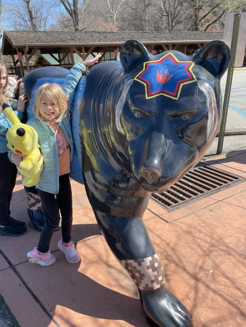A girl poses next to a large, colorful bear statue, holding a yellow stuffed animal, with trees in the background.