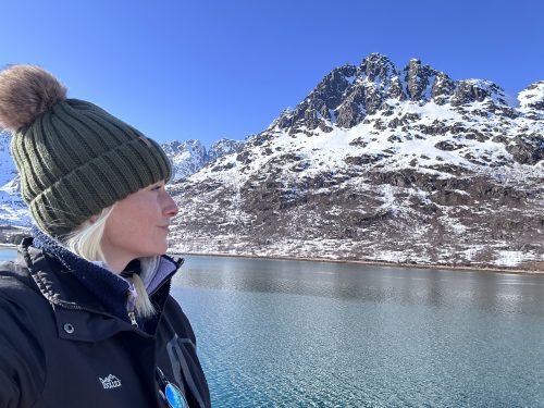 A woman in a green beanie gazes at snow-capped mountains by a calm, blue lake under a clear sky.