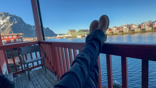 A person relaxes on a porch with feet up, overlooking a calm waterway and colorful houses against a mountain backdrop.