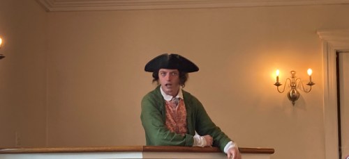 A man in historical attire, including a tricorn hat, stands at a balcony, looking surprised or animated.