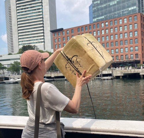 A woman in a cap holds a large, wrapped package over the edge of a boat, with buildings and water in the background.