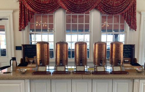 Five beverage dispensers on a counter, with a coffee pot and signs in a bright room with large windows.