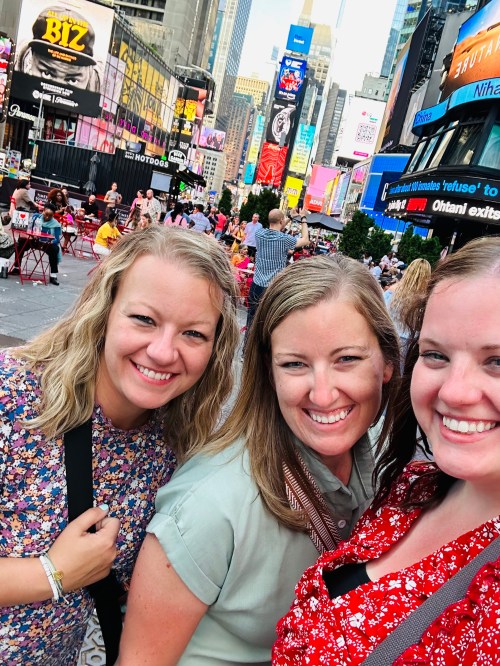 Three smiling women pose together in Times Square, surrounded by bright billboards and a bustling crowd.