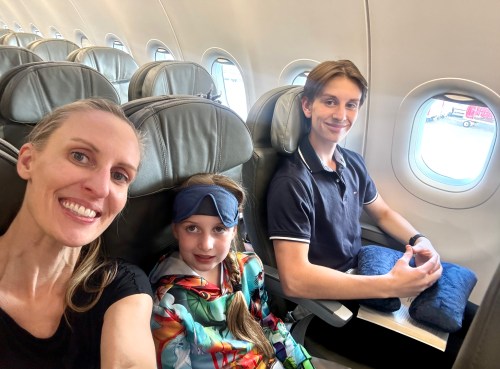 A woman and two children smile for a selfie on an airplane, with empty seats and a window in the background.
