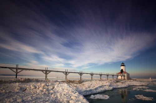 A lighthouse stands on a snowy shore, with a wooden walkway extending over icy waters under a dramatic sky.
