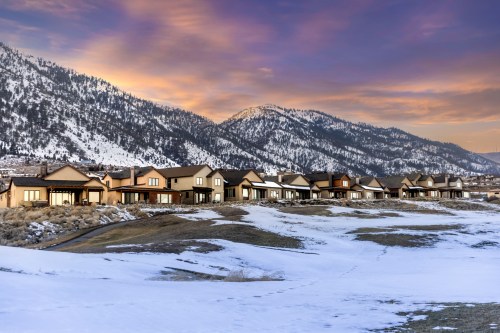 A row of houses on snowy ground with mountains and a colorful sky in the background at sunset.
