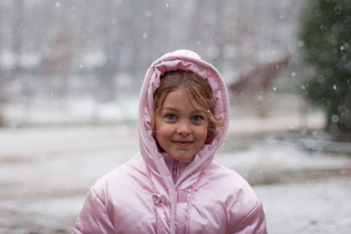 A young girl in a pink coat smiles warmly as snowflakes fall around her.